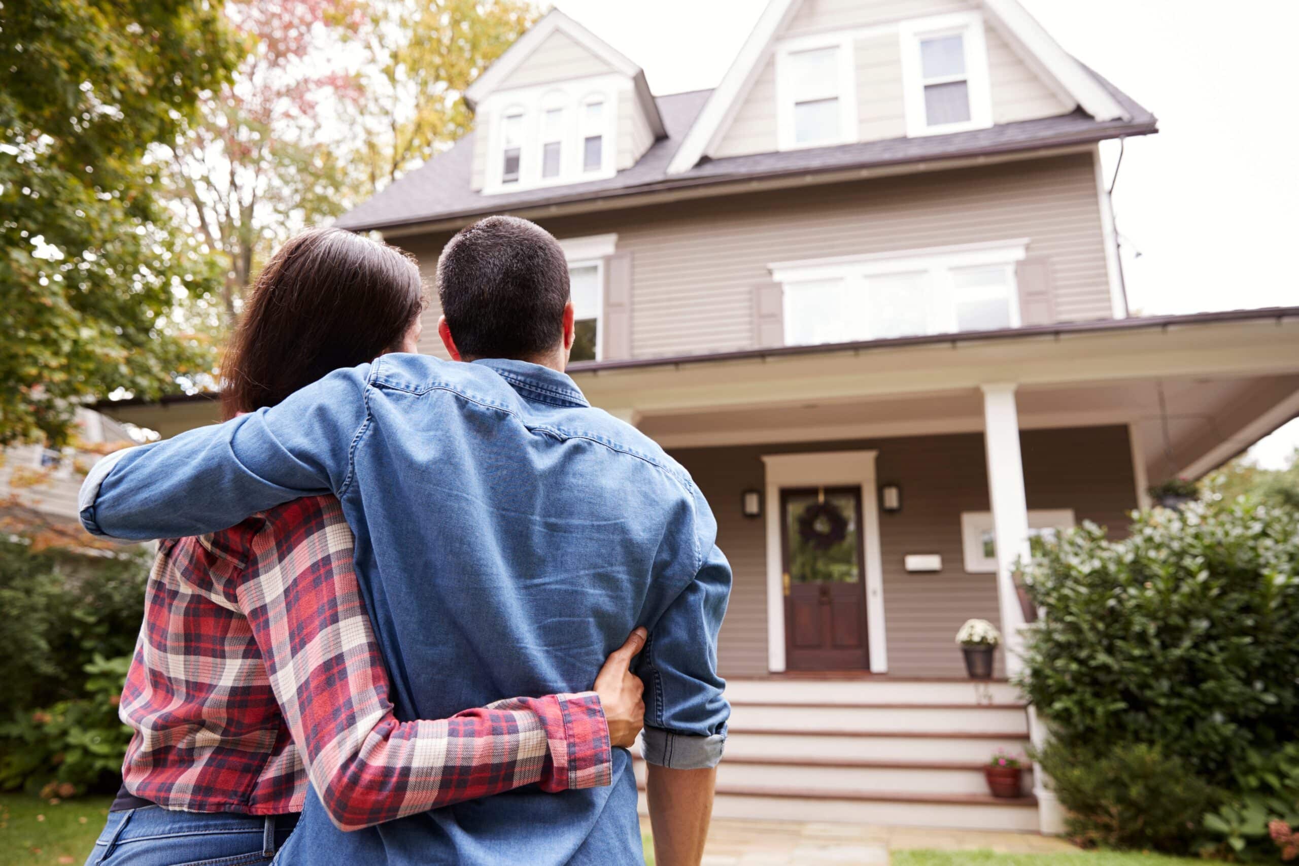 Couple embraces while viewing their new two-story home surrounded by greenery, symbolizing homeownership dreams.