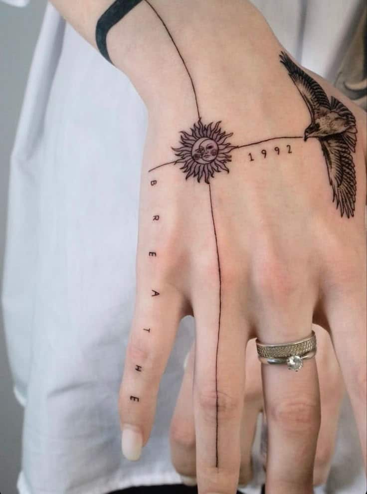 Hand tattoo with sun, bird, and BREATHE text, featuring the year 1992 and jewelry on the middle finger.