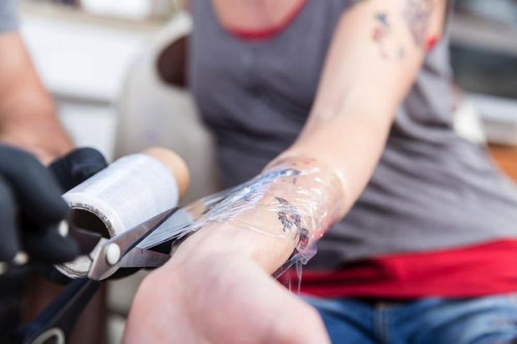 Person's arm with fresh tattoo being wrapped in protective plastic film by artist.