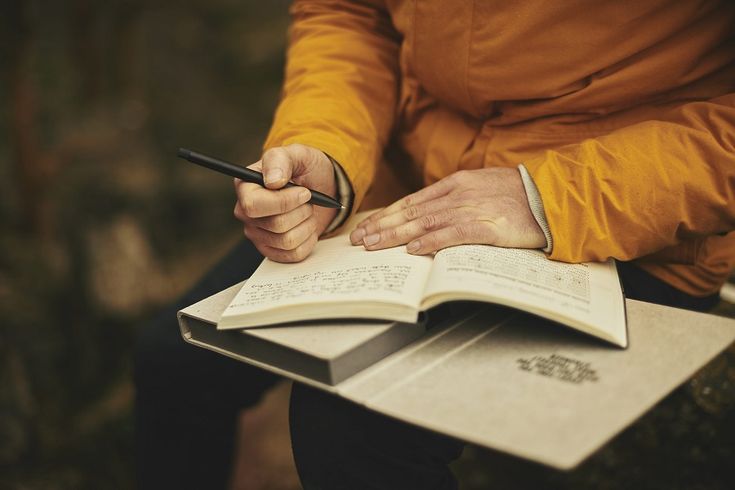 Person in orange jacket writing in journal outdoors, focusing on creative ideas and reflection.