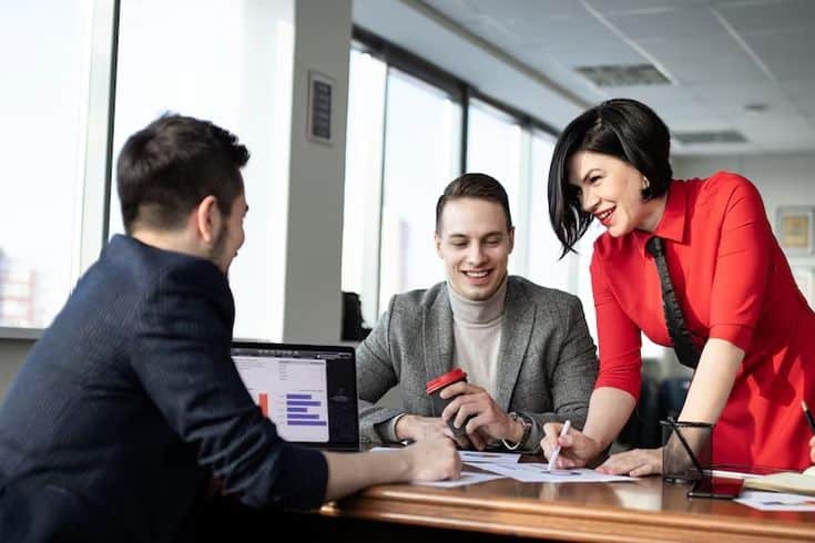 Three colleagues in a bright office collaborate around a table with documents and a laptop displaying graphs.