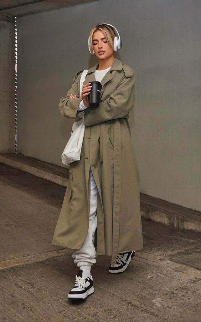 Woman in a trench coat and headphones walking, holding a coffee cup and wearing sneakers. Urban fashion style.