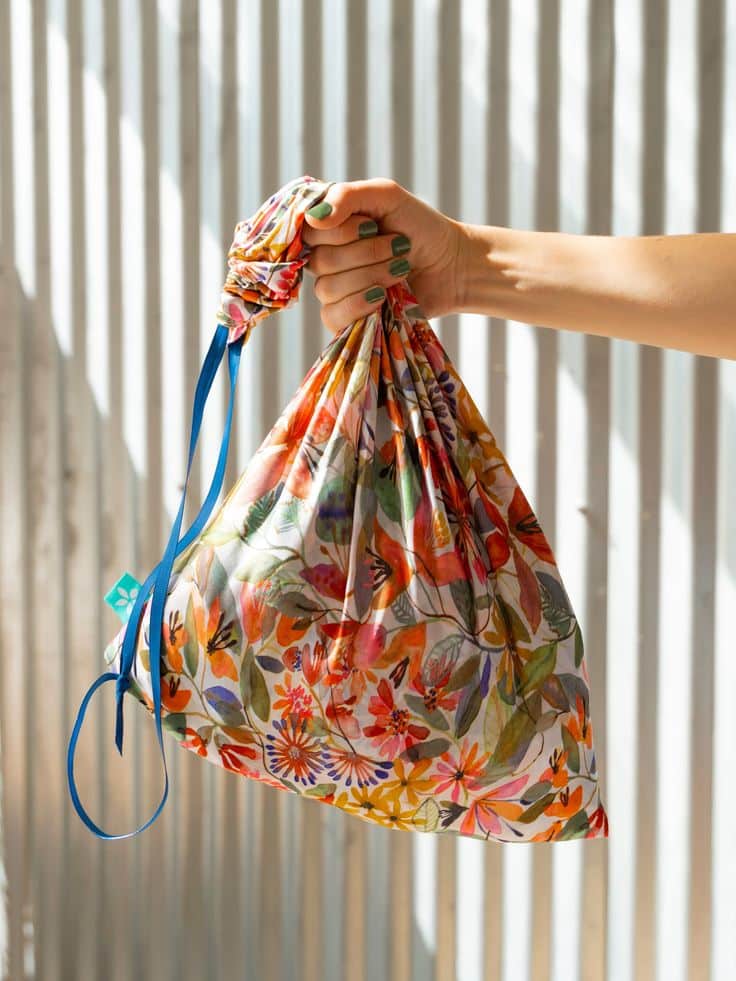 Hand holding a reusable floral bag with blue string, in front of sunlight and striped background. Eco-friendly shopping.