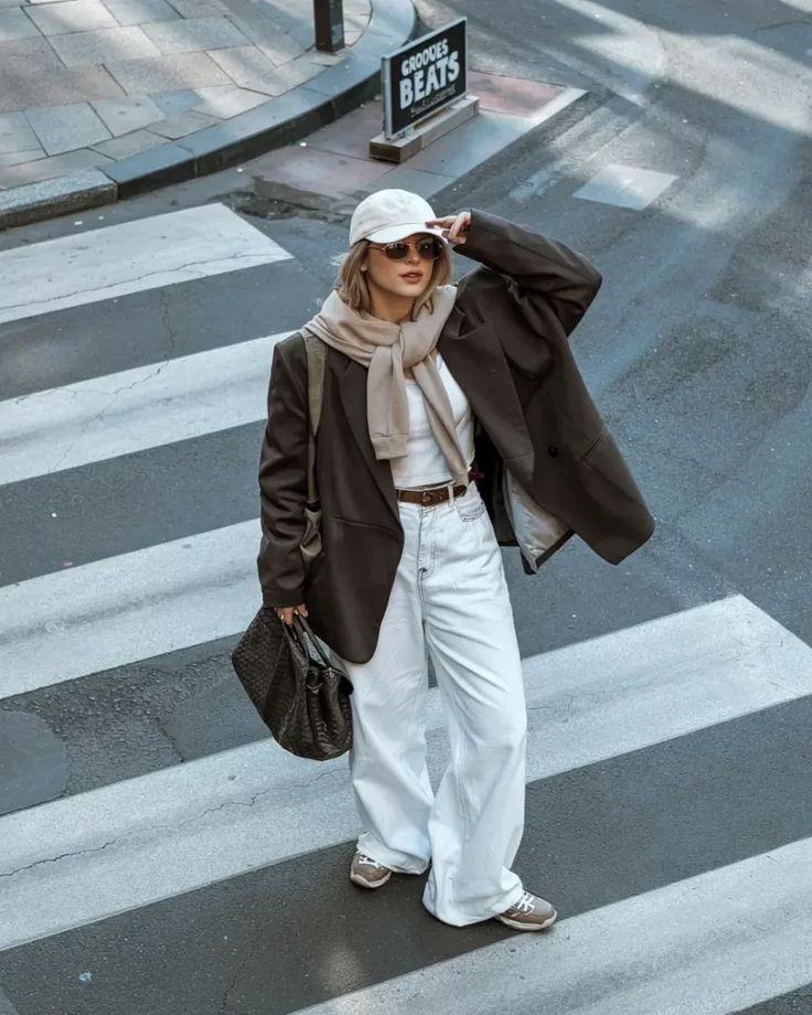 Fashionable woman crossing street in chic blazer, wide-leg jeans, sneakers, and cap. Urban style outfit.