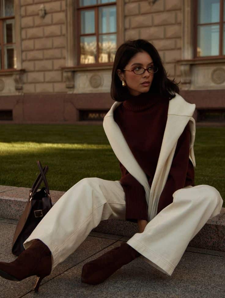 Stylish woman in chic fall outfit with glasses, seated near elegant building, showcasing autumn fashion trends.