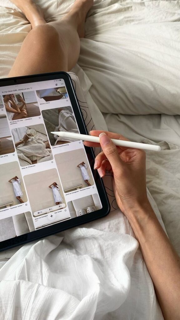 Person browsing fashion images on tablet in bed, holding a stylus.