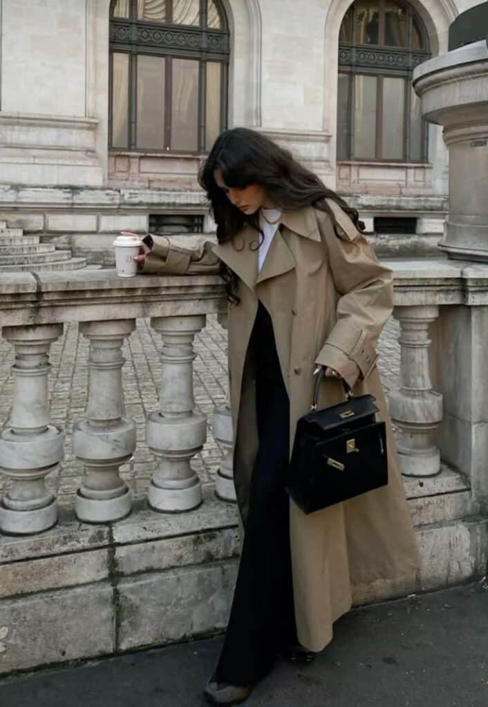 Woman wearing a beige trench coat holding coffee and black handbag in an urban setting.