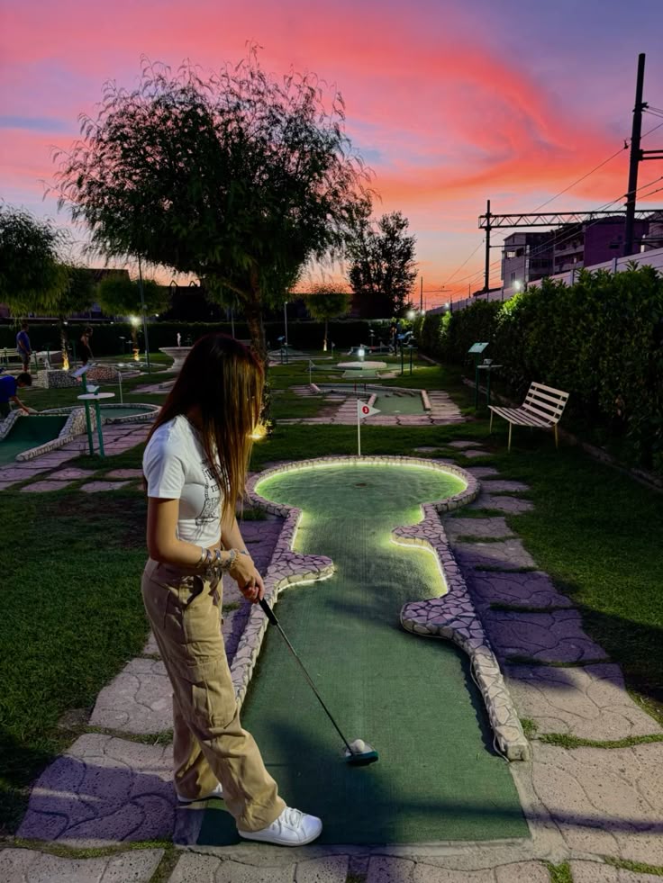 Person playing mini golf at sunset, colorful sky in the background, focused on putting the ball into a lit hole.