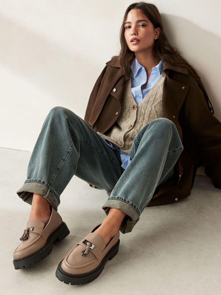 Casual fashion outfit with brown loafers, jeans, brown coat, blue shirt, and beige cardigan, sitting against a white wall.