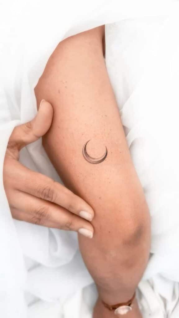 Minimalist crescent moon tattoo on a forearm with a hand gently touching, against a white fabric background.