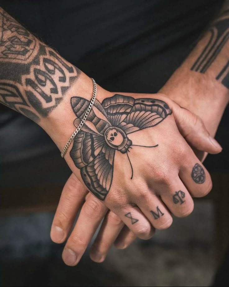 Hand tattoo featuring a detailed moth design with geometric symbols on fingers, and a chain bracelet on the wrist.
