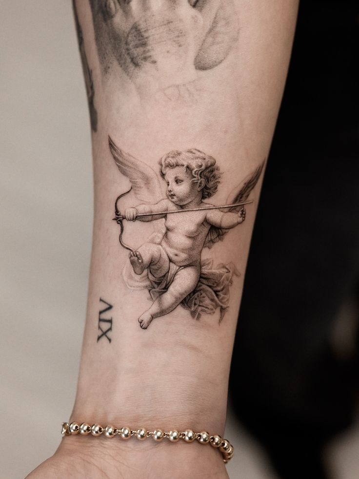 Never get bored working on a cherub tattoo👼🏻__blackandgreytattoo cherubtattoo angeltattoo cherub Angel tattoo on forearm with Roman numeral XIV, paired with a gold bead bracelet, on light skin. | Sky Rye Design Angel tattoo on forearm with Roman numeral XIV, paired with a gold bead bracelet, on light skin.