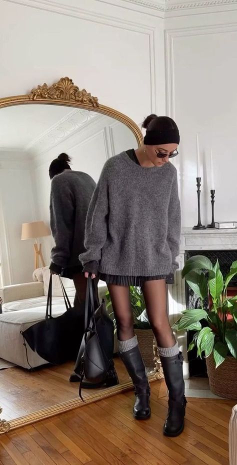 Fashionable woman in cozy gray sweater and boots, carrying bags in elegant room with mirror and plants.