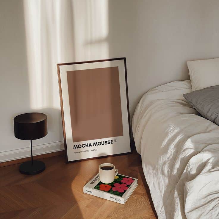 Framed art titled Mocha Mousse on floor next to bed, lamp, and book with coffee cup in a cozy, sunlit room.