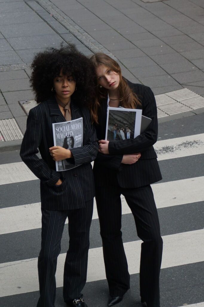 Two stylish women in pinstripe suits holding magazines, standing on a city street with a crosswalk.