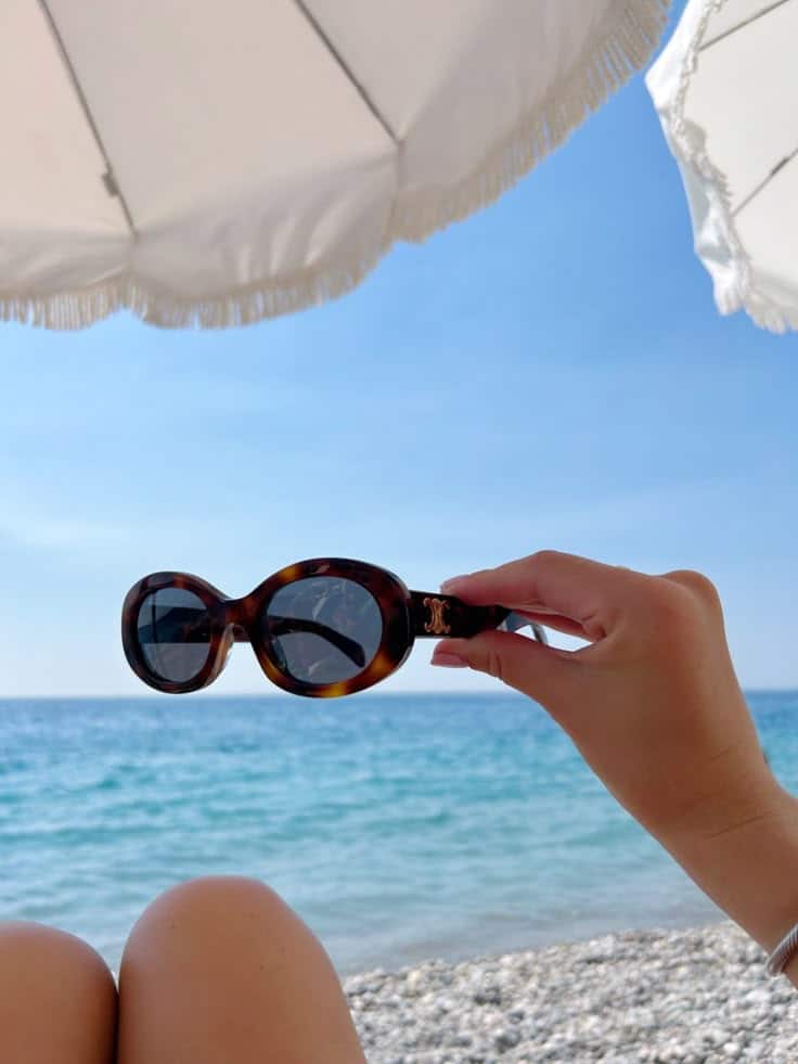 Sunglasses held under beach umbrella with ocean in background, relaxing summer day at the seaside.