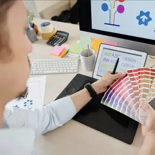 Designer selects color swatches at desk with tablet and computer displaying design software, enhancing creativity and workflow.