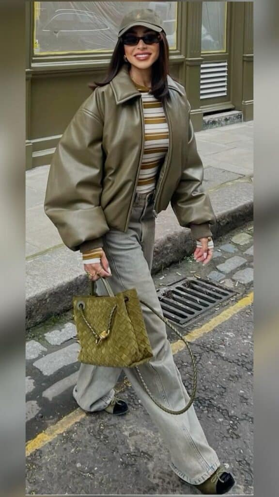 Fashionable woman in olive jacket and cap, striped shirt, gray jeans, holding woven bag, walking on a cobblestone street.