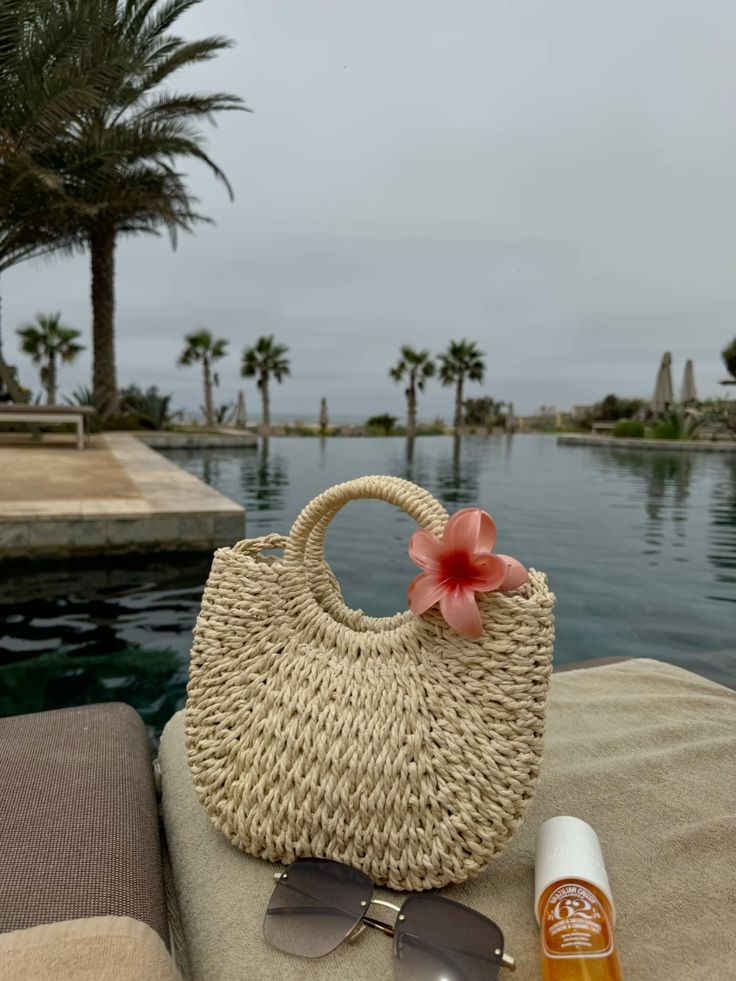Straw beach bag with pink flower, sunglasses, and sunscreen by a pool with palm trees in the background.