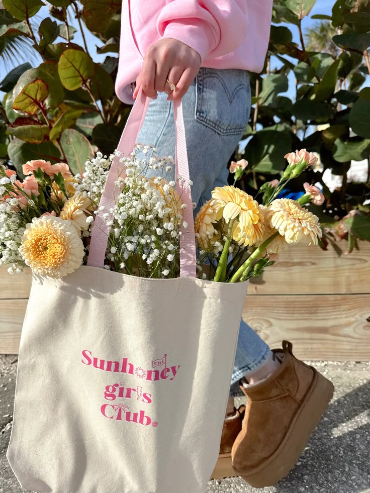 THE SUNHONEY GIRLS CLUB TOTE Person carrying a bag with flowers, wearing a pink sweater and jeans, outdoors. | Sky Rye Design Person carrying a bag with flowers, wearing a pink sweater and jeans, outdoors.