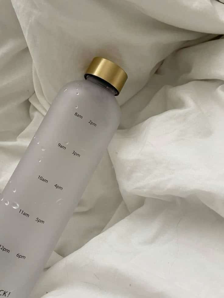 Frosted water bottle with gold cap on white sheets, marked with time indicators for tracking daily water intake.