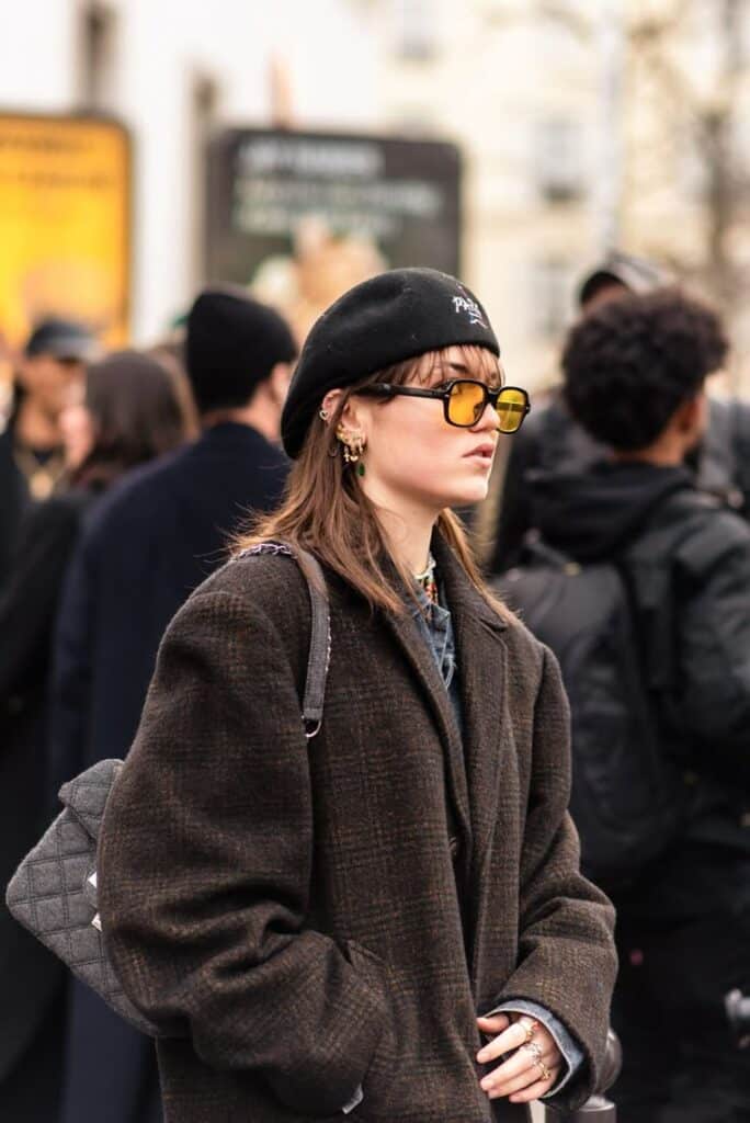 Fashionable person in brown coat and beret, wearing yellow sunglasses, with urban street style vibes at outdoor event.
