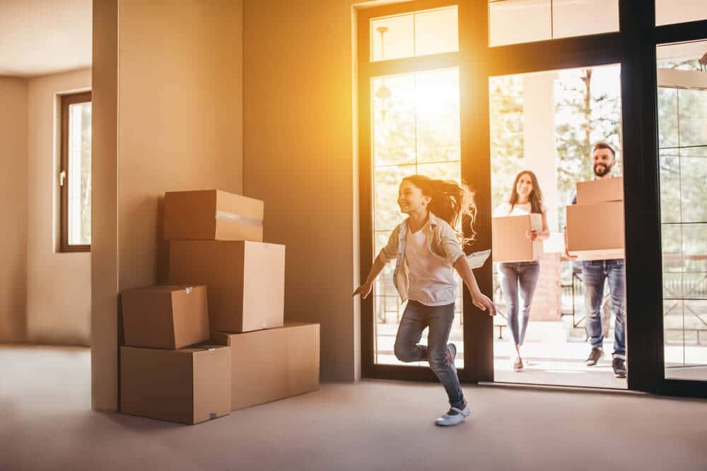 Family moving into new home with boxes, sunlight streaming through windows, and child joyfully running inside.