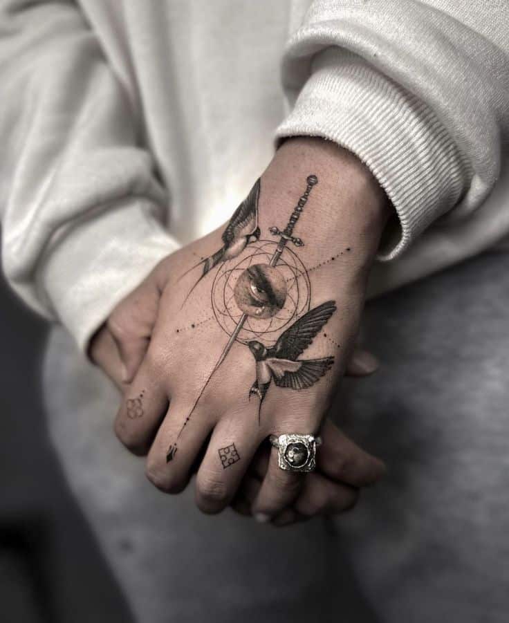 Close-up of a hand tattoo featuring a sword, birds, and an eye, paired with a silver ring and geometric symbols.