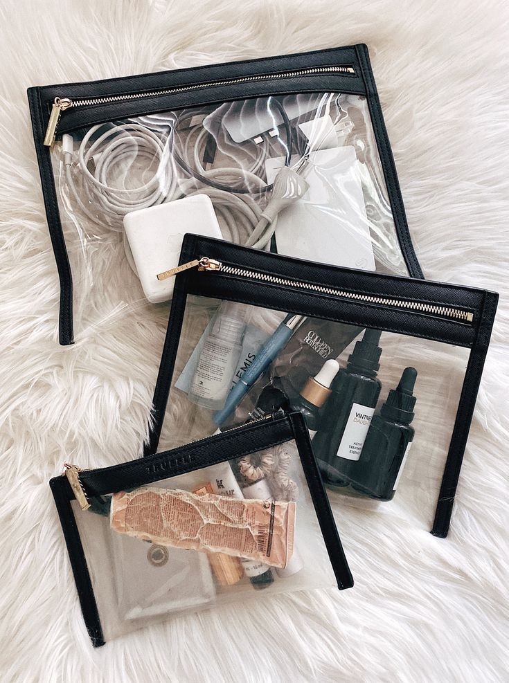 Clear pouches on soft white surface containing electronic accessories, skincare bottles, and toiletries for travel.
