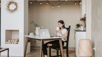 Modern home office with woman at a desk, using a laptop and sipping coffee, surrounded by cozy decor and soft lighting.