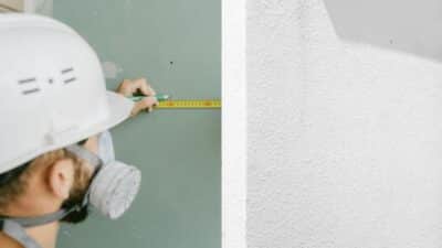 Construction worker measuring a wall with a tape measure, wearing a hard hat and mask for safety.