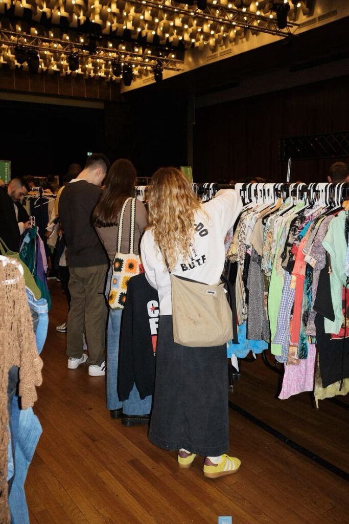 People browsing colorful vintage clothes at a thrift market under bright ceiling lights.
