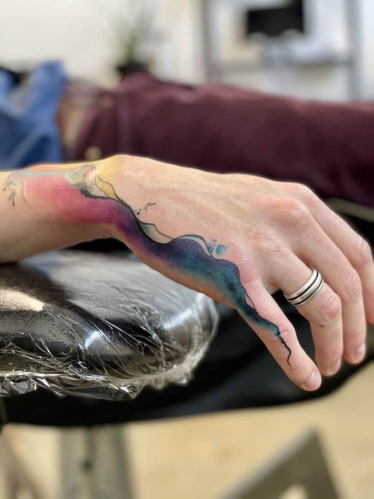 Colorful watercolor tattoo on arm, flowing abstract design, with rings on fingers and tattoo studio background.