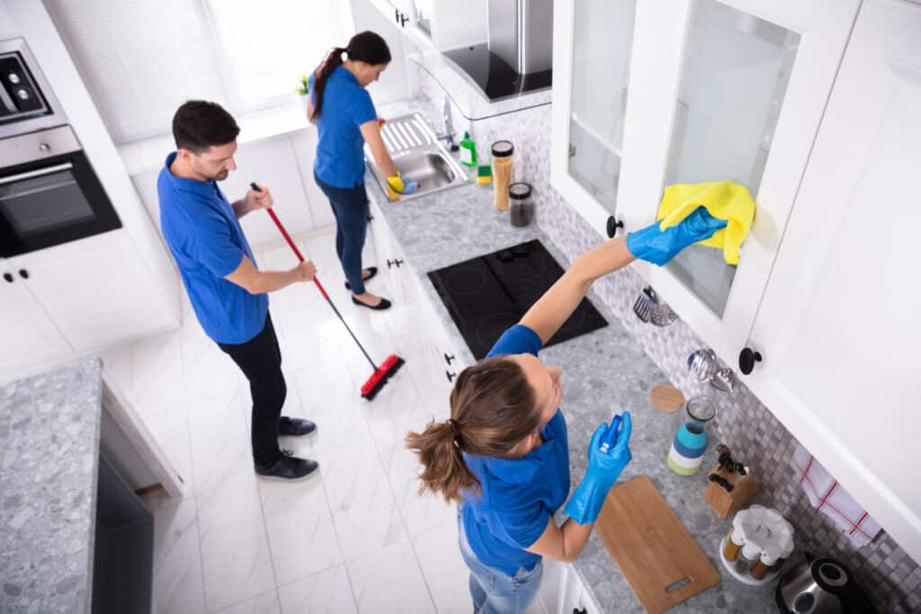 Three professional cleaners tidying a modern kitchen, wiping surfaces and sweeping the floor, wearing blue uniforms.