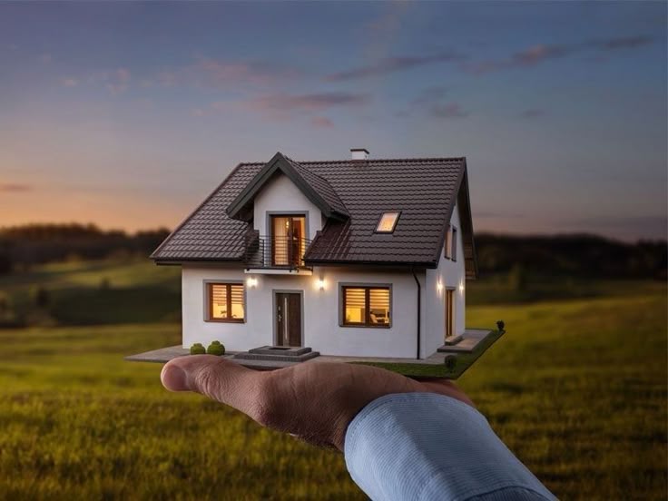 Hand holding a miniature house with lights glowing at dusk, symbolizing real estate investment and property ownership.