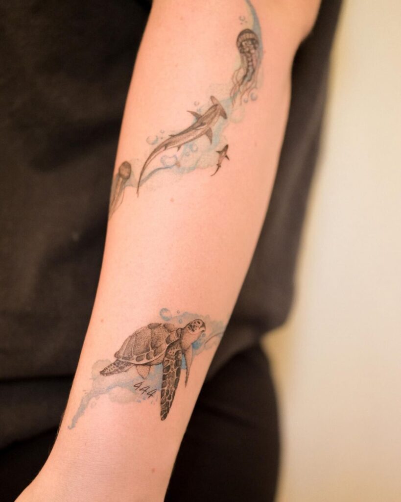 Tattoo of sea life on arm, featuring a turtle, shark, and jellyfish in watercolor style.