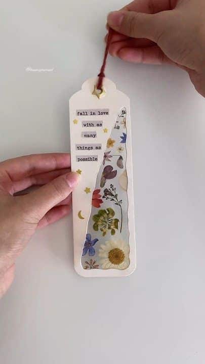 Hand holding a decorative bookmark with pressed flowers and quote: Fall in love with as many things as possible.