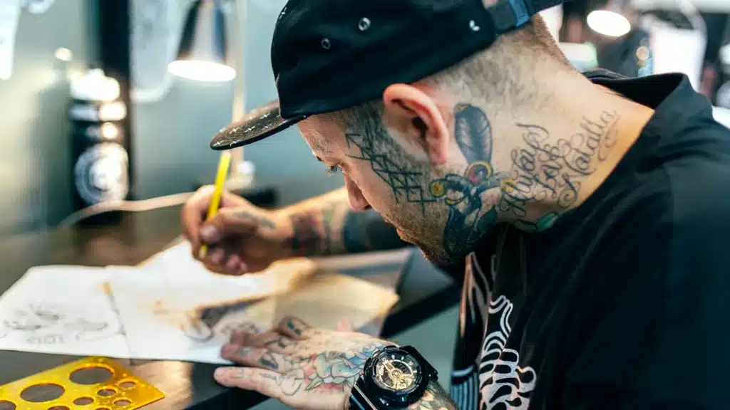 Tattoo artist sketching designs at a desk, surrounded by professional tools and sketches.