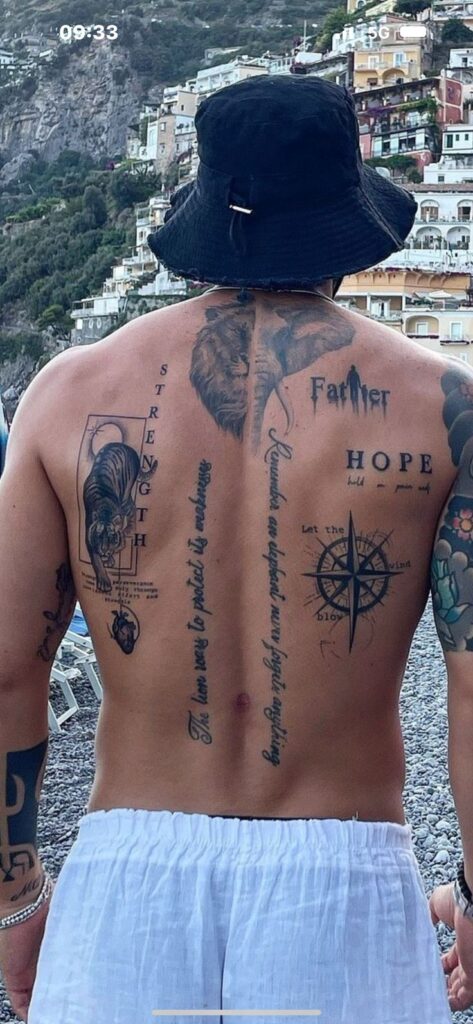 Man with tattoos and bucket hat facing a vibrant coastal village backdrop.