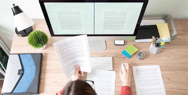 Person working at a tidy desk with computer, papers, and office supplies, focusing on document editing and organization.