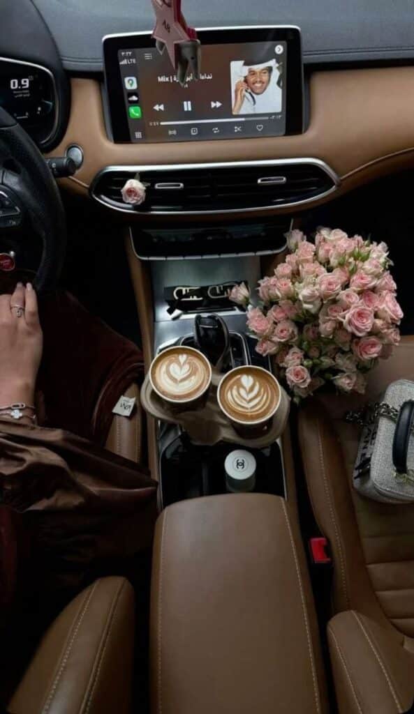 Car interior with cappuccinos, bouquet of pink roses, and music playing on dashboard screen for a cozy drive.