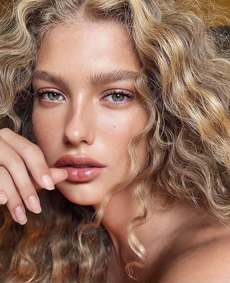Young person with curly blonde hair and natural makeup gazes thoughtfully, highlighting soft skin and elegant features.