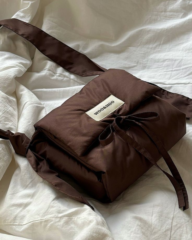 Brown padded shoulder bag with a bow, resting on white sheets, labeled Woohoo. Trendy fashion accessory.