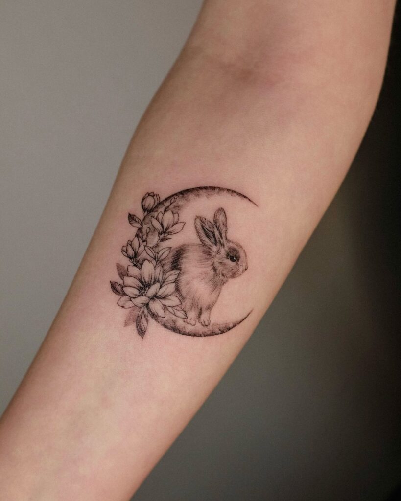 Tattoo with a bunny on a crescent moon surrounded by flowers, on a forearm.