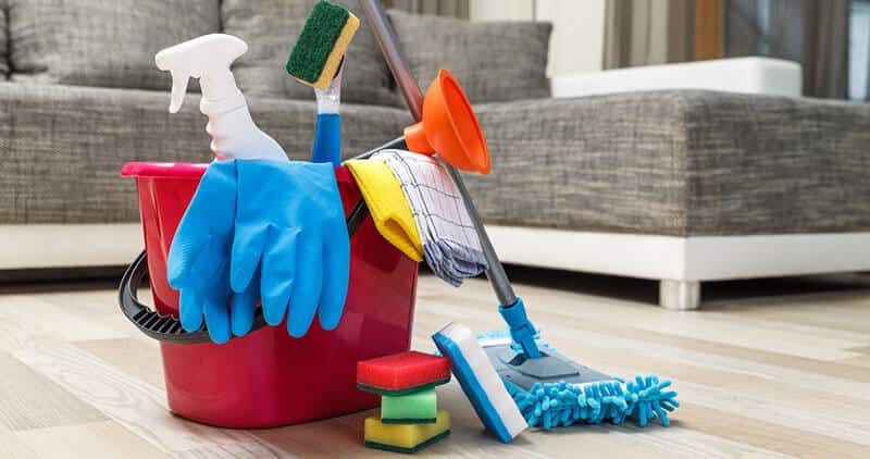 Cleaning supplies on wooden floor include bucket, gloves, sponges, mop, and spray bottle in a modern living room setting.