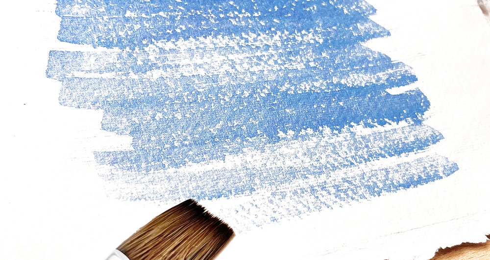 Paintbrush creating blue watercolor texture strokes on white paper, showcasing artistic technique and gradient effect.
