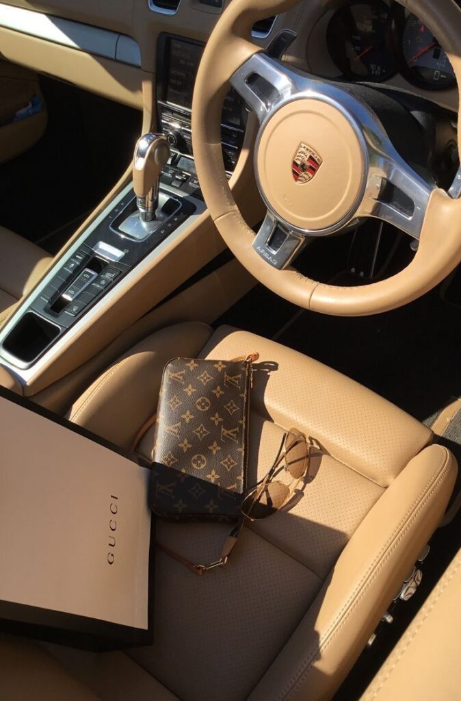 Luxury car interior with designer handbag, Gucci bag, and sunglasses on tan leather seat.