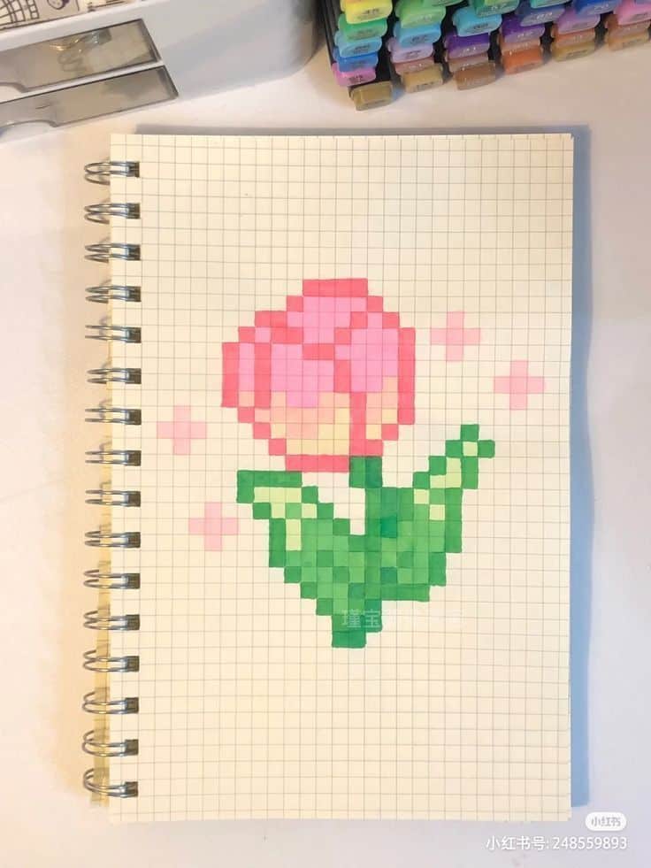 Pixel art tulip drawing on a grid notebook page, surrounded by colorful markers. Ideal for DIY and crafting inspiration.