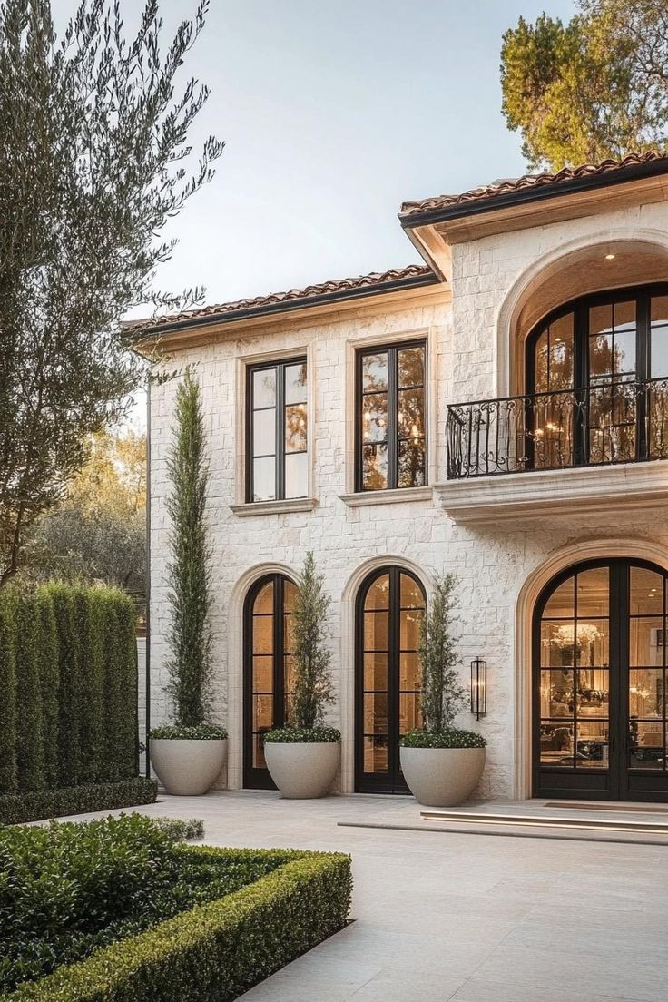 Elegant stone house with arched windows, tall trees in planters, and manicured hedges under a clear sky.