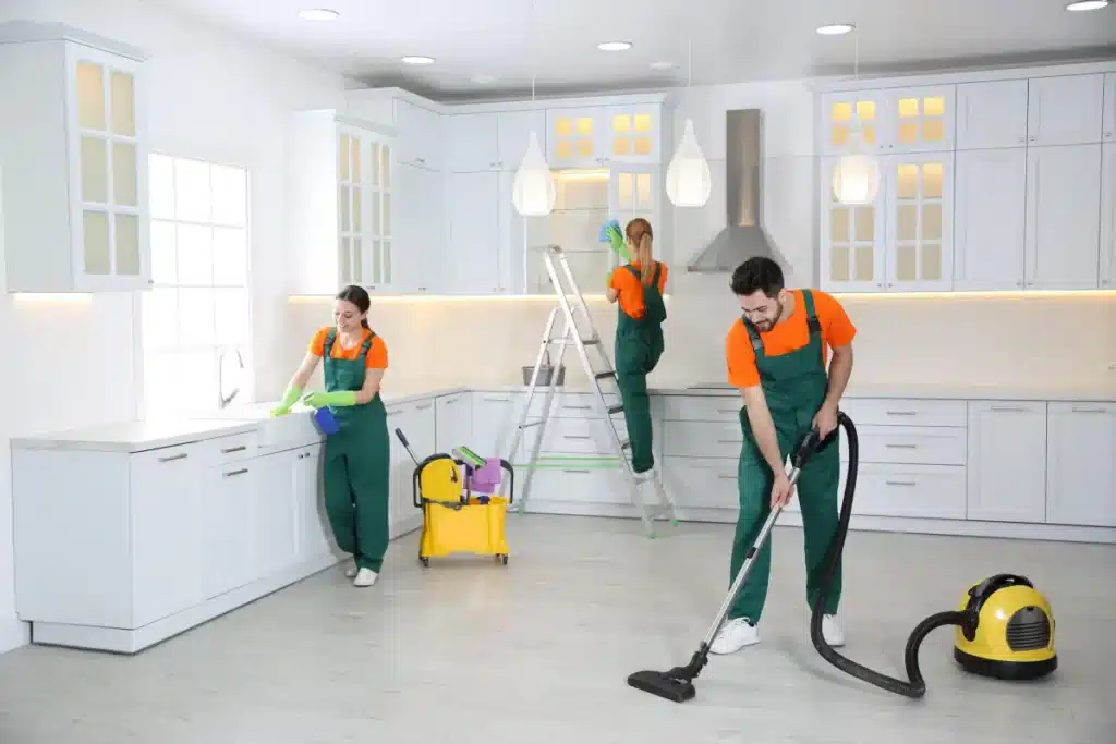 Cleaning crew in uniform tidying a modern kitchen with vacuum, mop, and wiping surfaces. Bright and organized space.